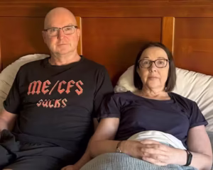 Peter McCluskey, carer for his wife Jenny, sitting up in bed. Peter wearing an "ME/CFS Sucks" t-shirt.